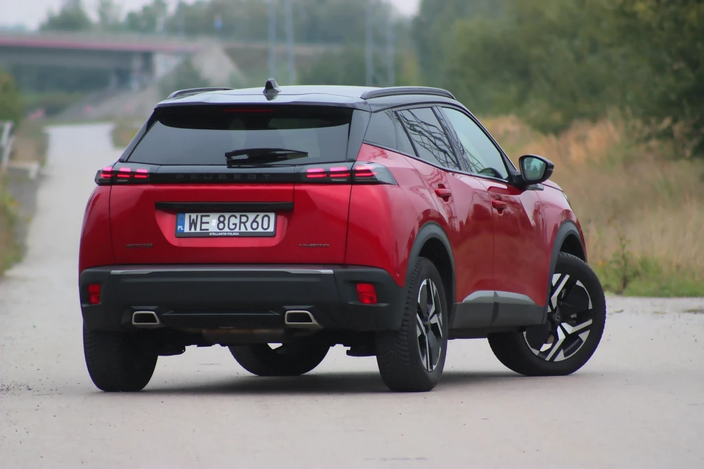 Mimo upływu 6 lat od premiery Peugeot 2008 wciąż wygląda nowocześnie Czerwony nowoczesny SUV zaparkowany na żwirowej drodze otoczonej zielenią, widok z tyłu pokazuje charakterystyczne światła oraz polską tablicę rejestracyjną.