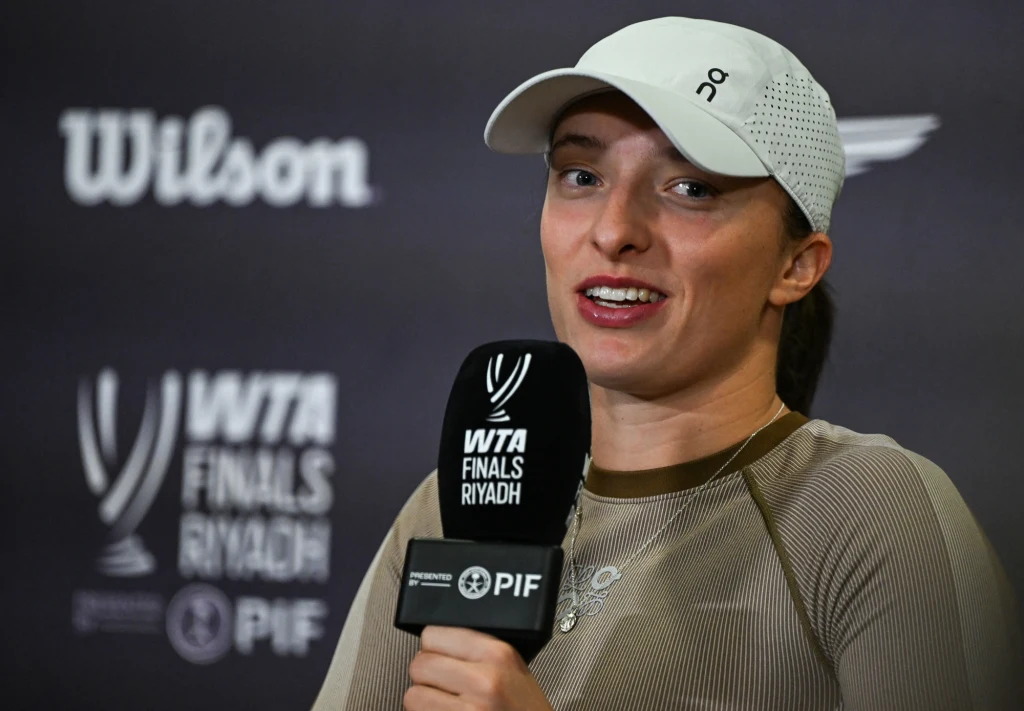 Iga Świątek Kobieta w sportowej czapce i koszulce trzyma mikrofon z logotypem turnieju tenisowego WTA Finals Riyadh, na tle ścianki z logo sponsorów wydarzenia.