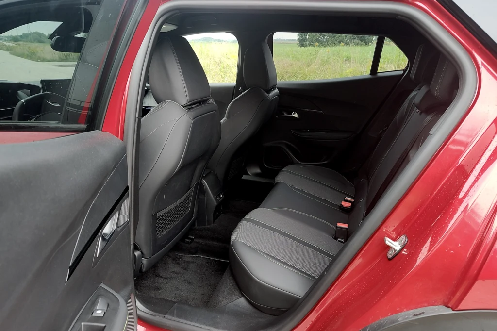 Ilość miejsca na tylnej kanapie Peugeota 2008 pozytywnie zaskakuje Tylna kanapa samochodu osobowego z uchylonymi drzwiami, trzy czarne siedzenia ze skórzanym obiciem, widok na zagłówek przedniego siedzenia i przestrzeń na nogi pasażerów.