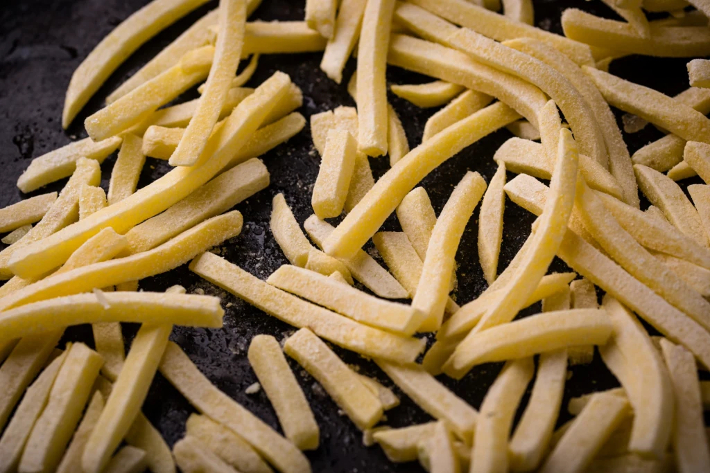 Rozsypane na ciemnej powierzchni zamrożone frytki, wyraźnie widoczne kryształki lodu pokrywające ich powierzchnię.