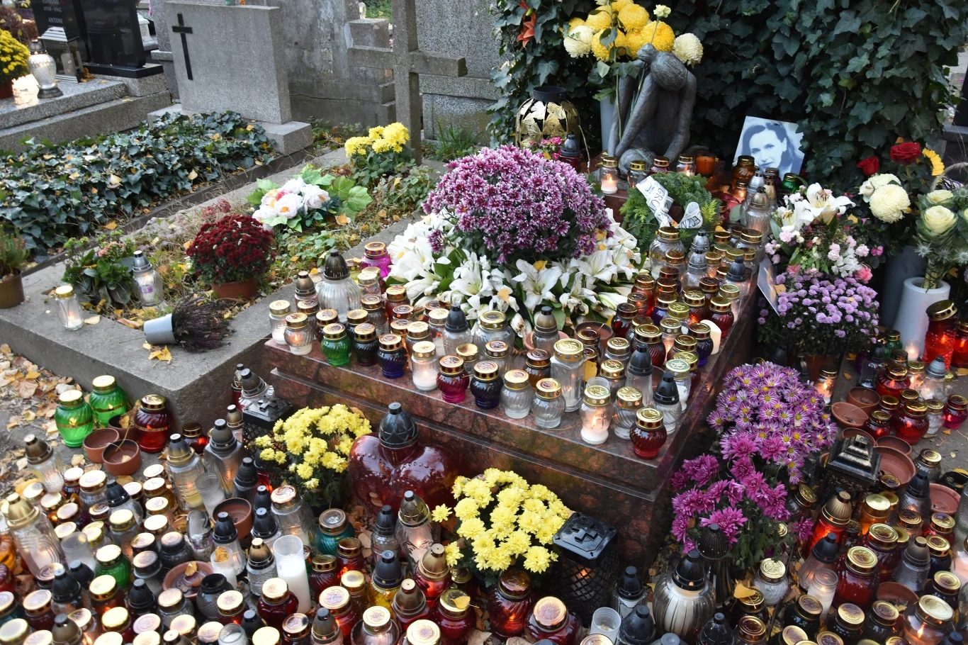 Tak wygląda grób Kory na Starych Powązkach w Warszawie - zdjęcie z 1 listopada 2024 r. Grób pokryty dużą ilością zapalonych zniczy, otoczony bukietami kwiatów, głównie chryzantem, oraz zdjęciem osoby zmarłej. W tle widoczne inne pomniki nagrobne i krzyże.