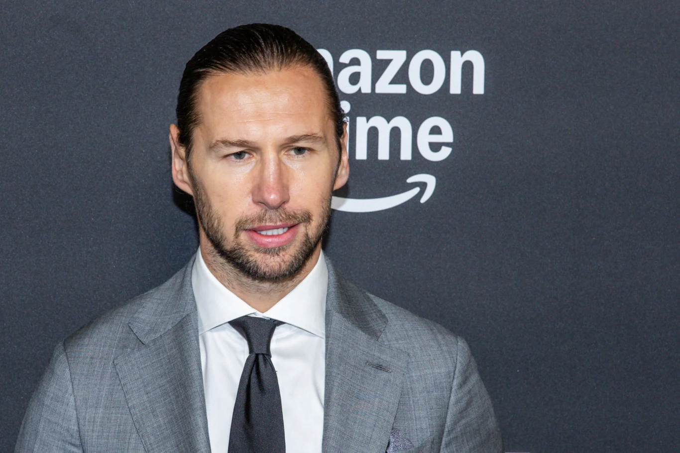 Mężczyzna w garniturze z ciemnym krawatem na tle czarnej ściany z logo Amazon Prime.