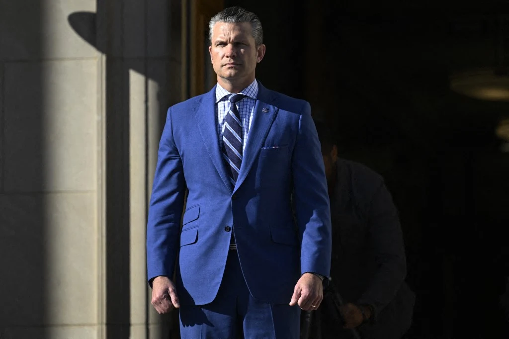 Sekretarz obrony USA Pete Hegseth Mężczyzna w dopasowanym, niebieskim garniturze i krawacie w jasne paski stoi przed wejściem do kamiennego budynku. W tle ciemne wnętrze, światło pada na postać z lewej strony.