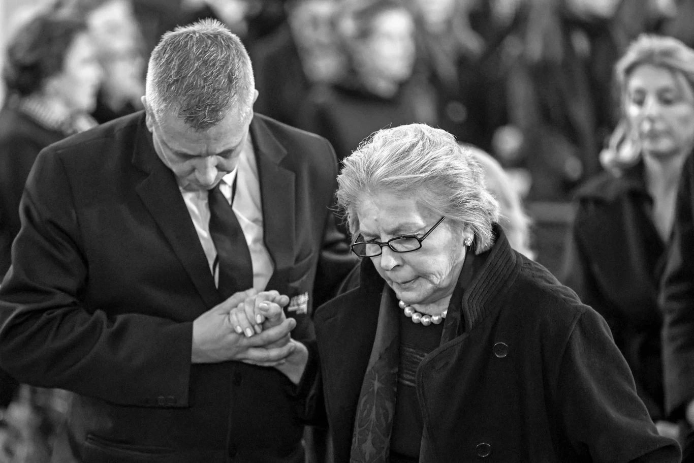 Elżbieta Penderecka miała 78 lat Starsza kobieta ubrana w ciemny płaszcz i perły prowadzona przez mężczyznę w garniturze, obydwoje wykazują powagę i skupienie, w tle stoją zamazane sylwetki innych osób.