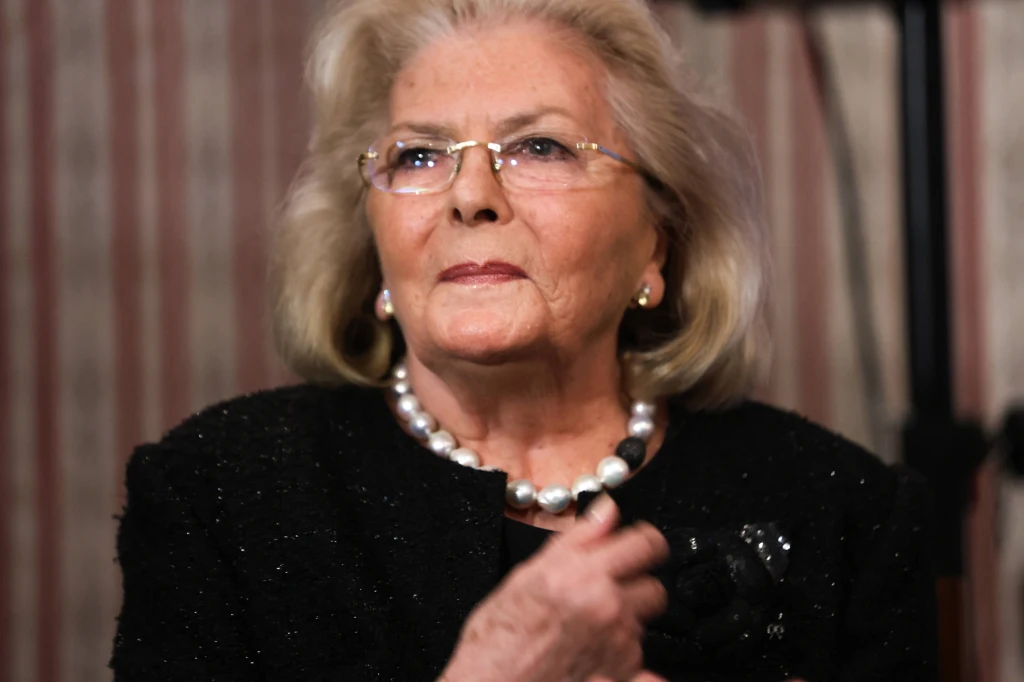 Starsza kobieta z jasnymi włosami, w okularach i eleganckiej ciemnej marynarce, nosi naszyjnik z dużych pereł i subtelne kolczyki. Tło z pionowymi pasami i elementami w ciemnych barwach.