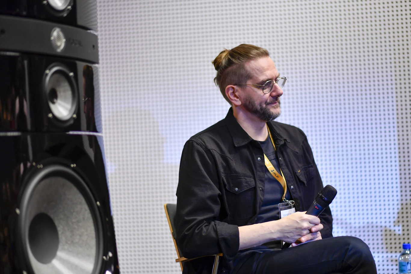 Lunatic Soul to projekt Mariusza Dudy Mężczyzna siedzący na krześle, trzymający mikrofon, ubrany w ciemną koszulę z plakietką na szyi, w tle duży głośnik oraz perforowana ściana.