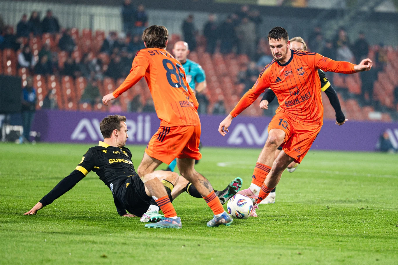 Mecz Bruk-Bet Termalica Nieciecza - GKS Katowice Dwóch piłkarzy w pomarańczowych strojach mierzy się z zawodnikiem w czarno-żółtej koszulce, który wykonuje wślizg, próbując odebrać piłkę. Na drugim planie widać kibiców na stadionie oraz sędziego obserwującego sytuację.