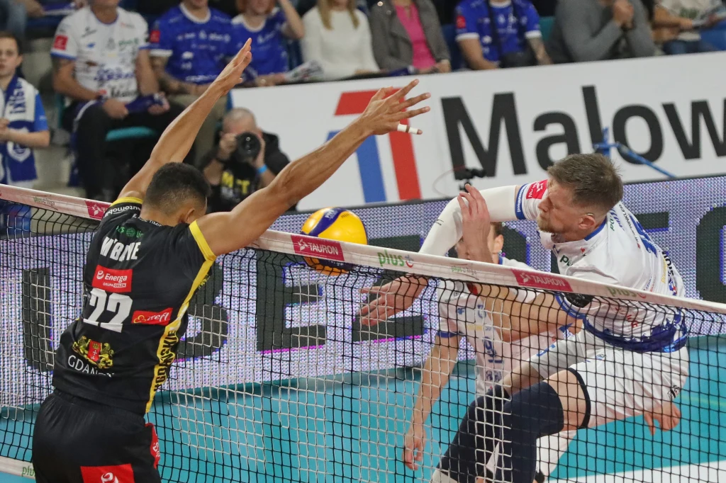 Kadr z meczu Ślepsk Malow Suwałki - Energa Trefl Gdańsk Dynamiczna akcja podczas meczu siatkówki, dwaj zawodnicy rywalizują przy siatce, jeden z nich wykonuje atak, drugi próbuje blokować piłkę. W tle widoczna jest publiczność oraz bandy reklamowe.