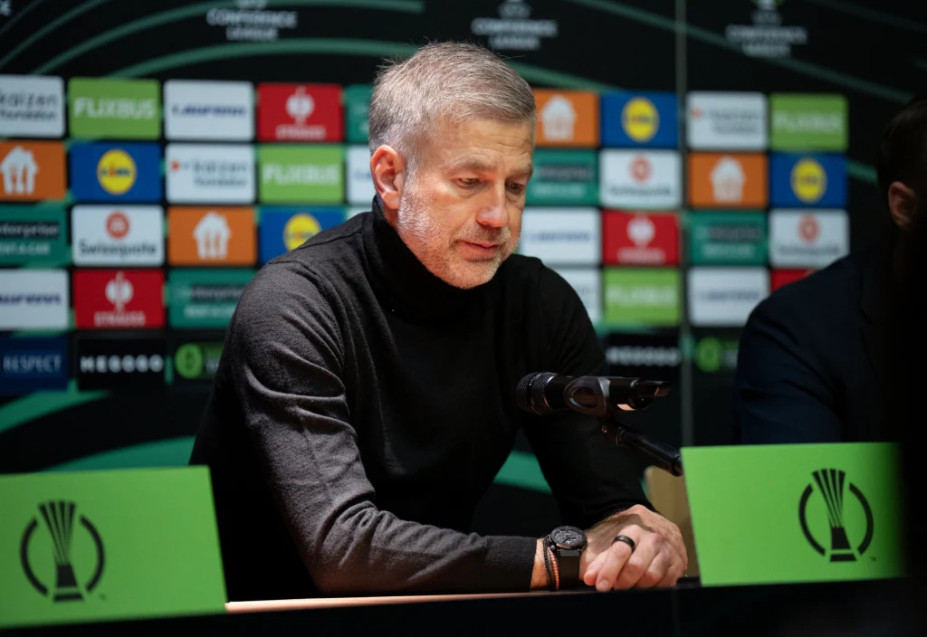 Mężczyzna siedzi za stołem konferencyjnym podczas konferencji prasowej, przed nim znajduje się mikrofon, a w tle widoczna jest ściana z logotypami sponsorów i organizatorów.