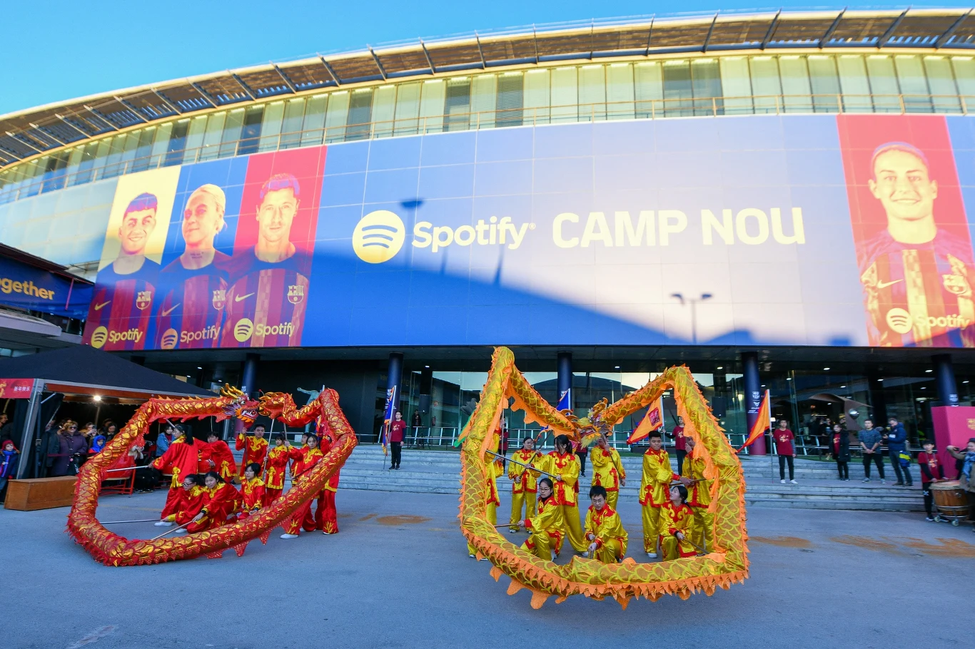 Spotify Camp Nou Tancerze w strojach nawiązujących do chińskiego stylu smoka wykonują taniec przed głównym wejściem na stadion Spotify Camp Nou, w tle ogromny baner z wizerunkami piłkarzy FC Barcelony.