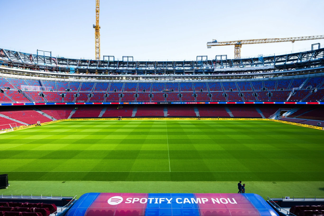 Spotify Camp Nou Pusta murawa stadionu piłkarskiego otoczona trybunami, z widocznym napisem 'Spotify Camp Nou' na pierwszym planie, w tle żurawie budowlane świadczące o prowadzonych pracach modernizacyjnych.