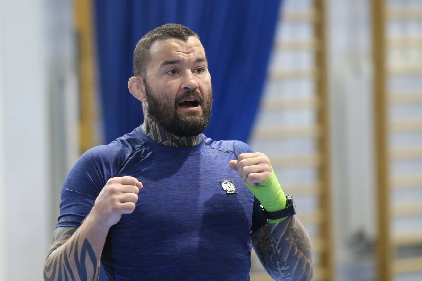 Mężczyzna ubrany w sportową koszulkę z tatuażami na ramionach i szyi, z opaską na lewej dłoni, znajdujący się w jasnym pomieszczeniu sportowym, gestykulujący jak podczas treningu.