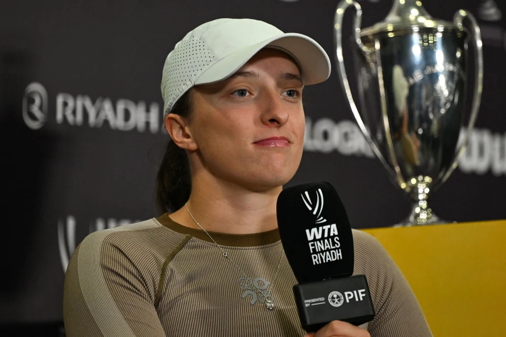 Iga Świątek na konferencji prasowej przed WTA Finals 2025 Kobieta w sportowej czapce trzyma mikrofon z logo WTA Finals Riyadh, za nią widoczny puchar turniejowy i fragment napisu z tła konferencji prasowej.