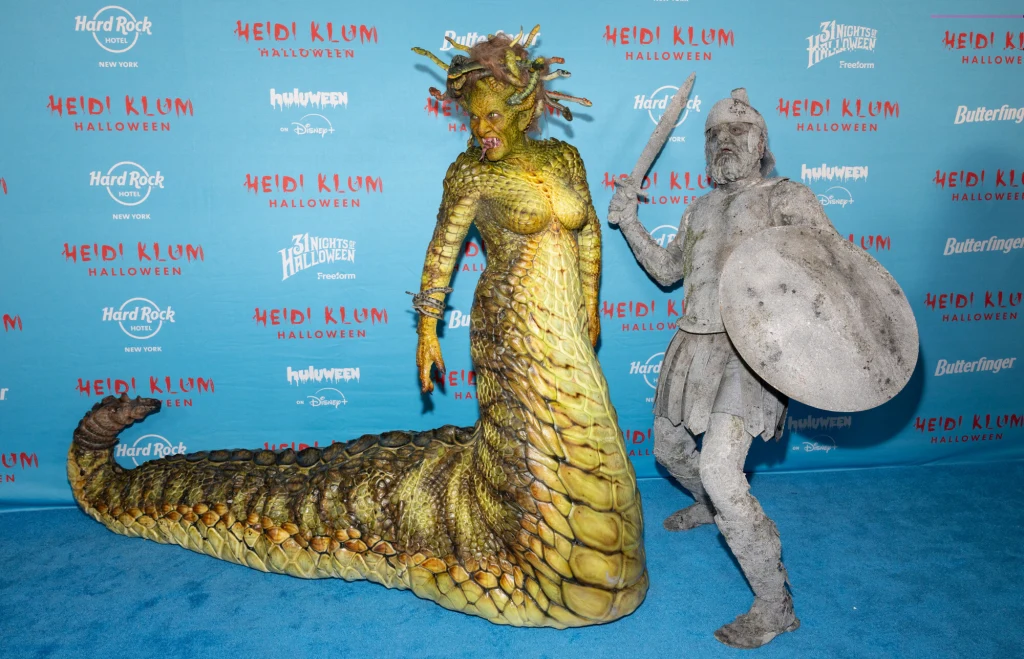 Heidi Klum i Tom Kaulitz Kobieta w imponującym kostiumie Meduzy z realistycznie wykonanym ogonem i wężami na głowie oraz mężczyzna przebrany za kamiennego wojownika z tarczą i mieczem, pozują razem na tle ścianki z logotypami wydarzeń Halloween.
