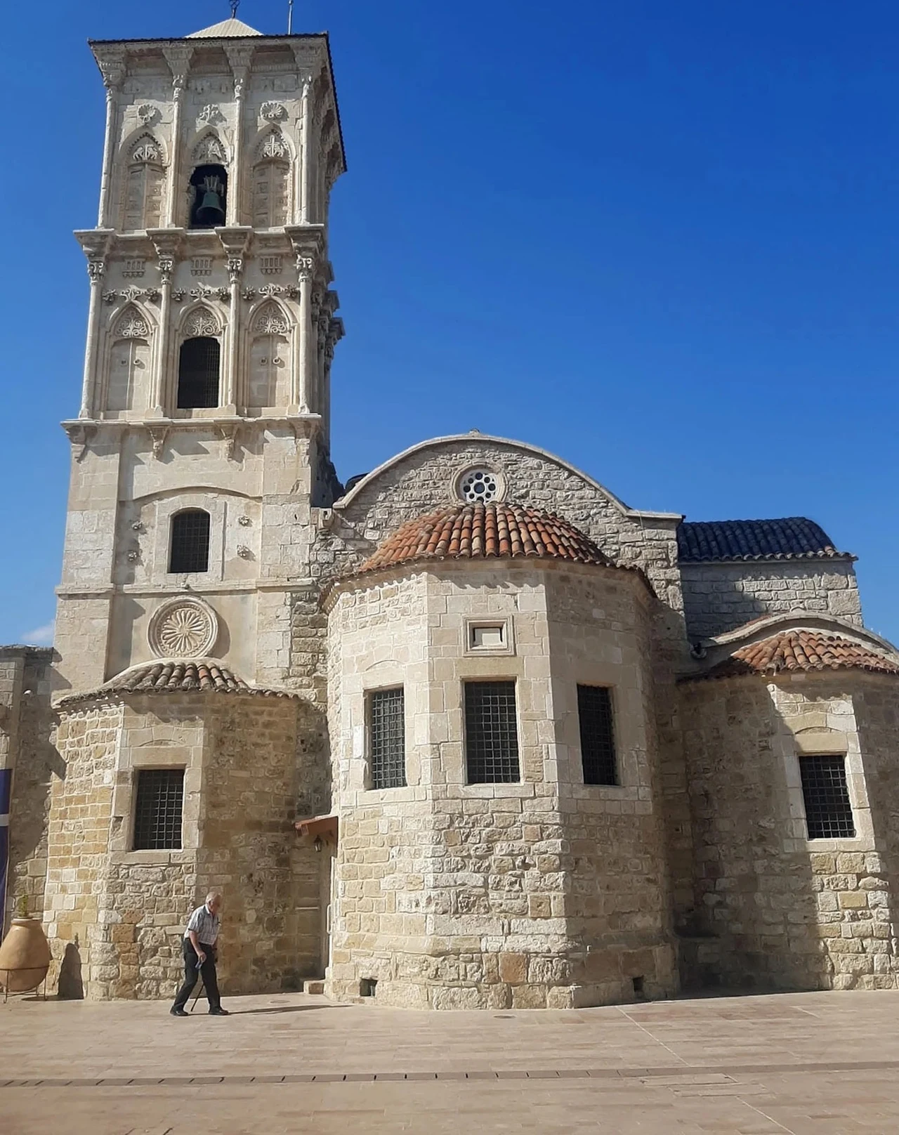 Cerkiew św. Łazarza w Larnace Kamienna cerkiew z wysoką wieżą dzwonniczą na tle błękitnego nieba, z boku przechadza się samotny mężczyzna. Budowla charakteryzuje się średniowieczną architekturą i zdobieniami wokół okien.