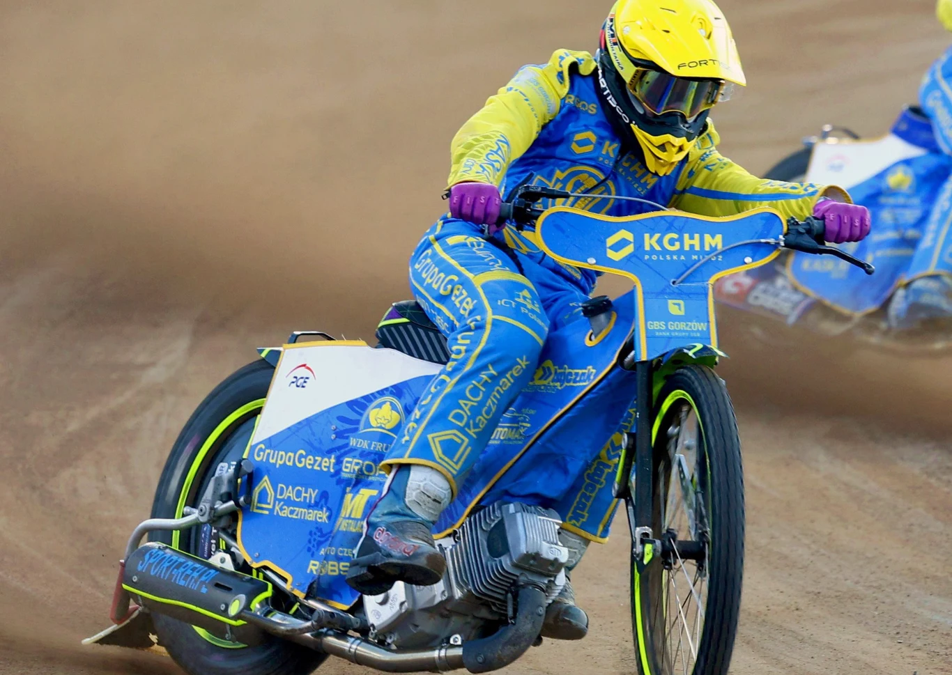 Zawodnik w pełnym ekwipunku żużlowym jedzie motocyklem po torze, unosząc za sobą kurz. Kask i kombinezon w jaskrawych kolorach, na motocyklu widoczne liczne logotypy sponsorów.