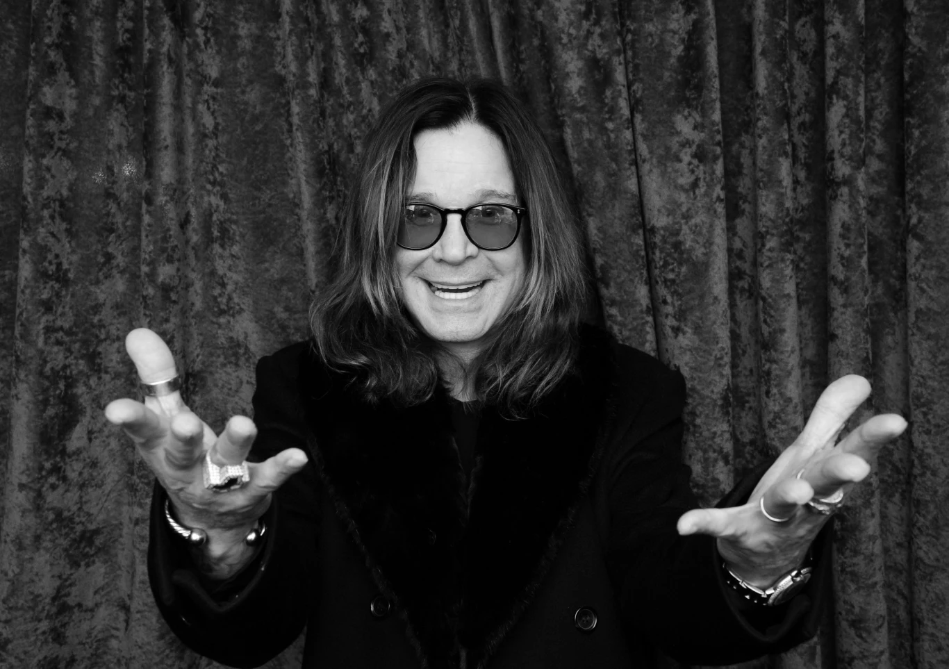 Ozzy Osbourne Uśmiechnięty mężczyzna w ciemnych okularach i długich włosach wyciąga ręce w stronę obiektywu. Na sobie ma ciemne ubranie oraz kilka pierścionków na palcach. Tło stanowi zasłona o welurowej fakturze.