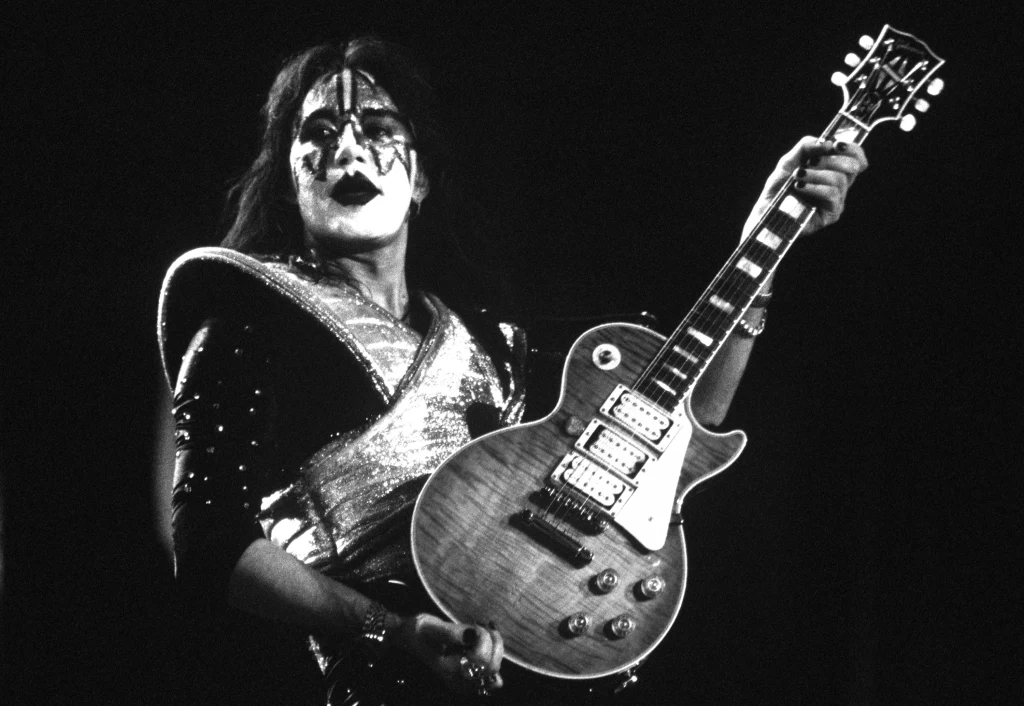 Muzyk w kostiumie scenicznym i makijażu charakterystycznym dla zespołu rockowego trzyma elektryczną gitarę, światło reflektorów podkreśla jego sylwetkę na ciemnym tle.