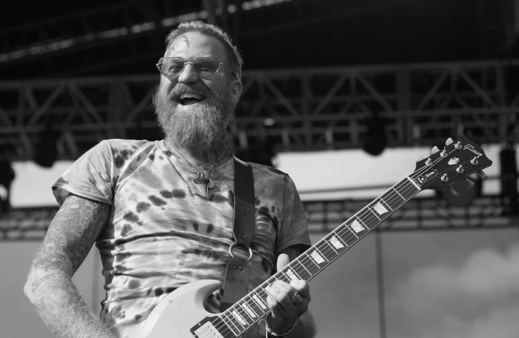 Brodaty mężczyzna w okularach przeciwsłonecznych, ubrany w koszulkę tie-dye, gra na gitarze elektrycznej na scenie plenerowej, uśmiechnięty, z widocznymi tatuażami na ramionach.