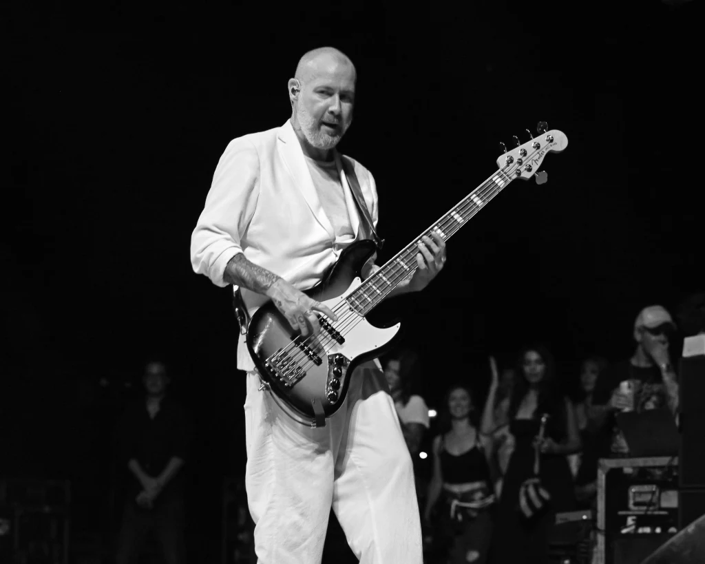 Mężczyzna grający na gitarze basowej na scenie koncertowej, ubrany w jasny garnitur, w tle widoczna grupa widzów oklaskujących występ.