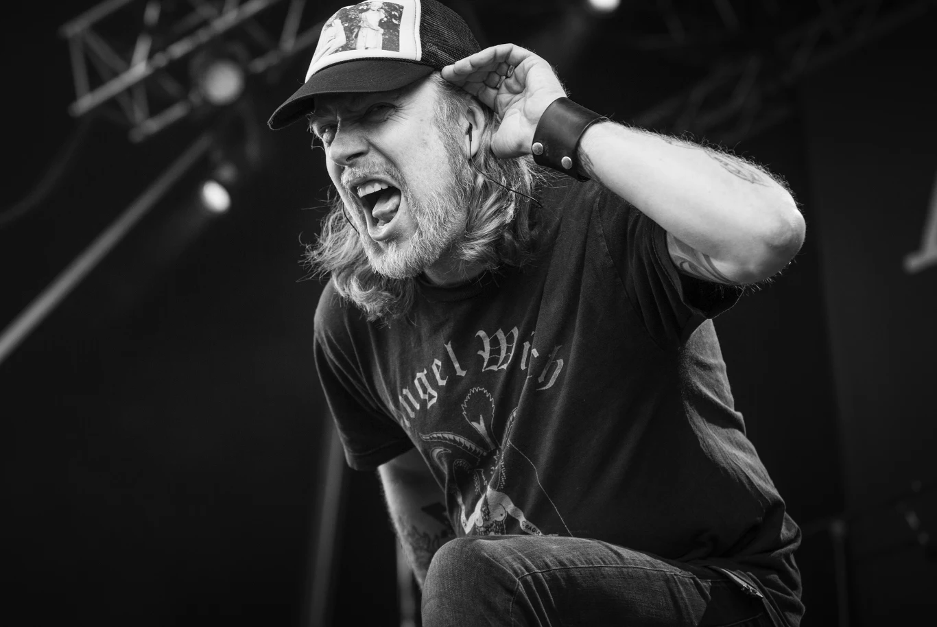 Tomas Lindberg (At The Gates) Mężczyzna z długimi włosami i brodą w czapce z daszkiem pochyla się do przodu, śpiewając lub krzycząc na scenie muzycznej, przykładając dłoń do ucha, jakby nasłuchiwał reakcji publiczności. Na koszulce widnieje wyrazisty napis.