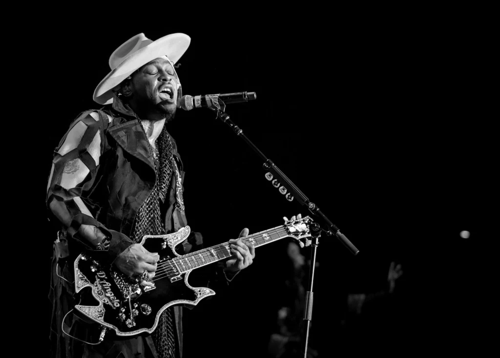 Mężczyzna w dużym kapeluszu śpiewa do mikrofonu, grając energicznie na gitarze elektrycznej, scena zanurzona w ciemnościach podkreślających ekspresję artysty.