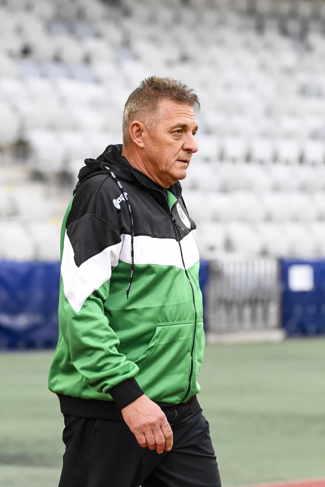 Mężczyzna w sportowej zielono-czarno-białej bluzie idący po stadionie, w tle rzędy pustych, jasnych krzeseł.