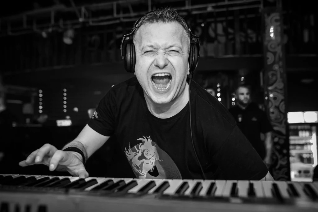 Mężczyzna w słuchawkach grający na klawiaturze elektronicznej, z szeroko otwartymi ustami w geście wyrażającym silne emocje, w tle rozmyte światła klubu i postać stojąca przy barze.