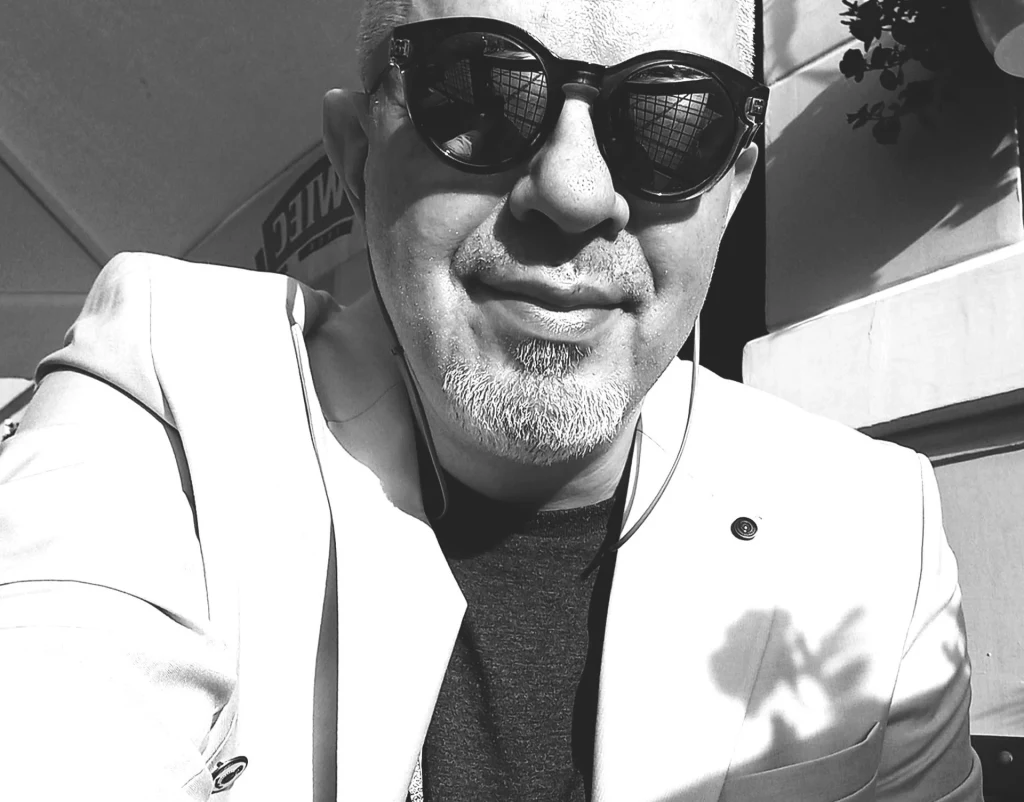 Mężczyzna w ciemnych okularach przeciwsłonecznych i jasnej marynarce patrzy wprost do obiektywu, siedząc na tle jasnej ściany w słoneczny dzień.