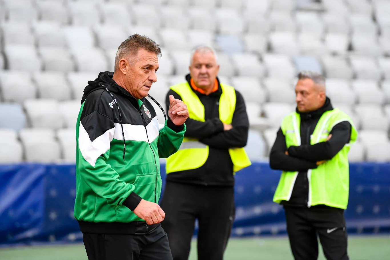 Trener w zielono-czarnej kurtce prowadzi rozmowę lub odprawę na stadionie, dwie inne osoby w żółtych kamizelkach asekuracyjnych słuchają go, siedzenia stadionowe w tle.