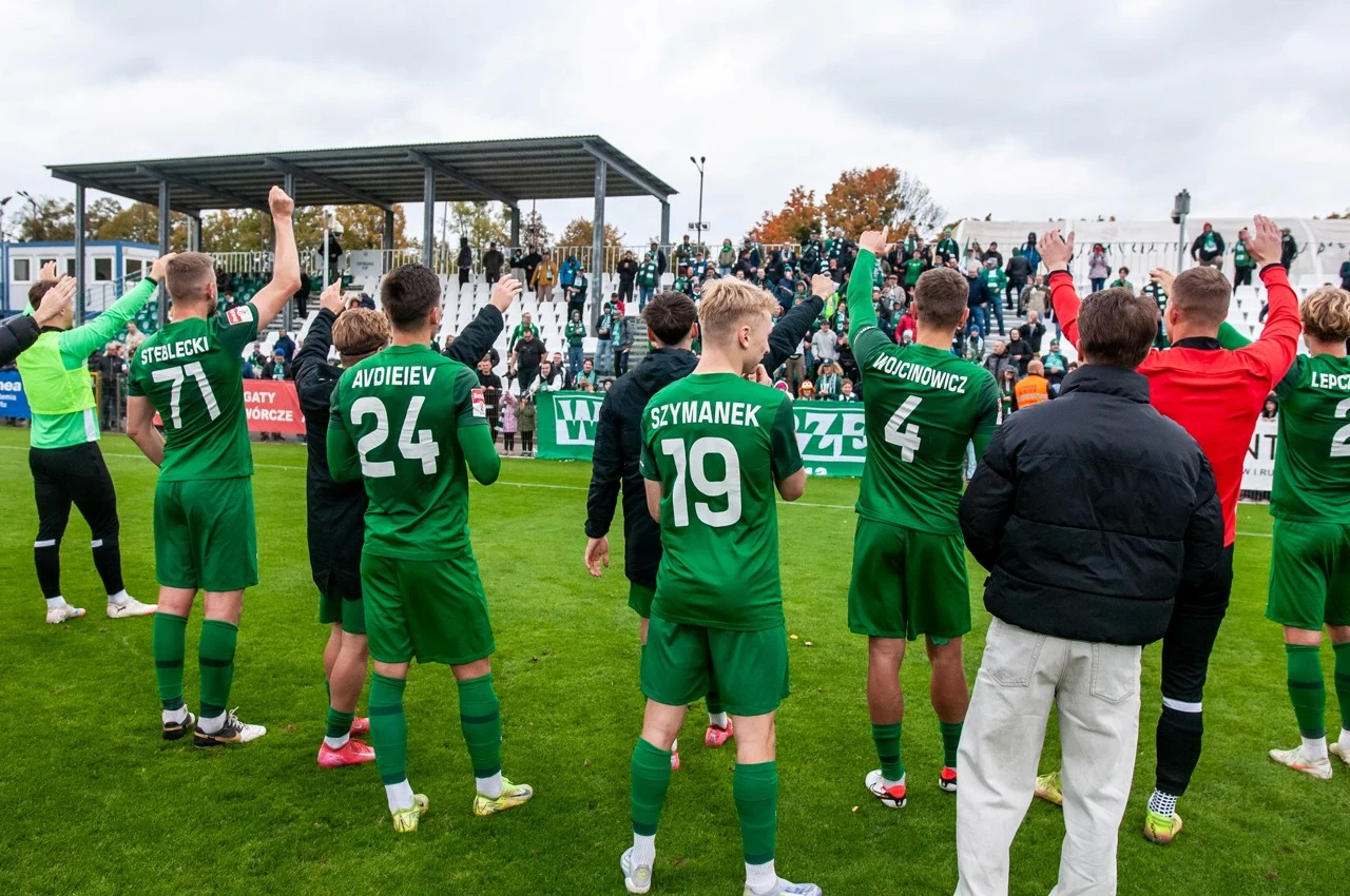 Grupa piłkarzy w zielonych strojach stoi na murawie stadionu oraz unosi ręce, kibicującym im osobom na trybunach. W tle widoczna jest zadaszona trybuna oraz liczna grupa kibiców odpowiadających gestami.