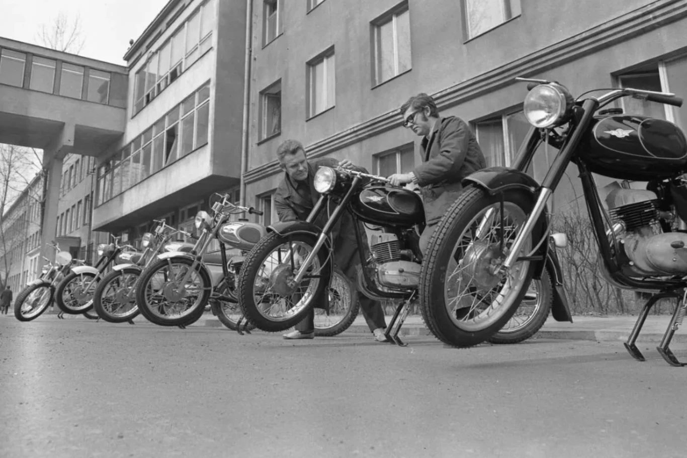 Ostatni motocykl marki WSK zjechał z linii produkcyjnej 31 października 1985 r. Szereg zabytkowych motocykli ustawionych w linii pod budynkiem, dwóch mężczyzn pochylonych nad jednym z motocykli, sprawdzającego jego stan techniczny.