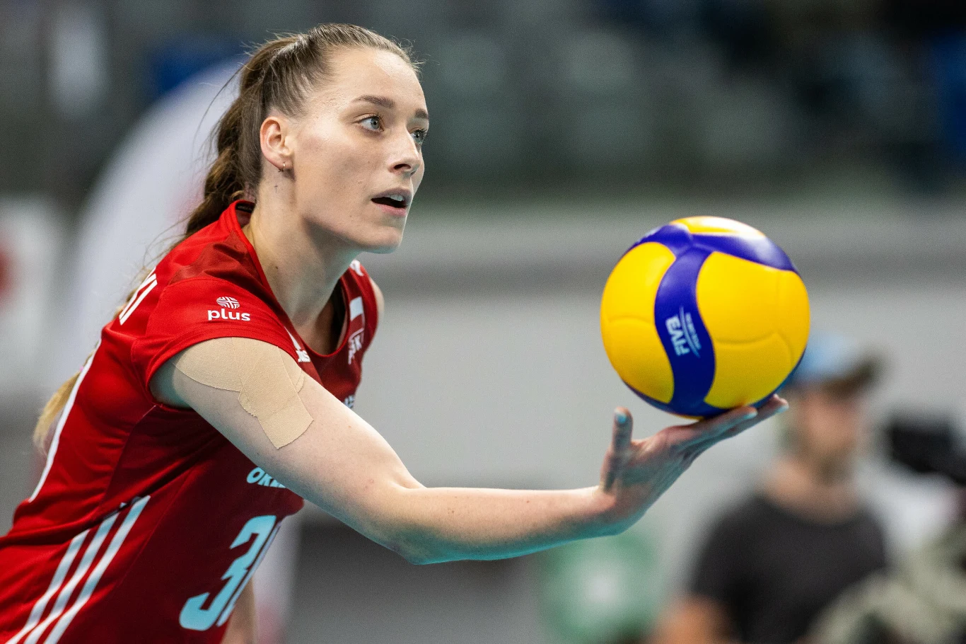 Siatkarka w czerwonym stroju z numerem 30 przygotowująca się do serwu, skupiona, trzymająca piłkę do siatkówki jedną ręką na tle rozmytego wnętrza hali sportowej.