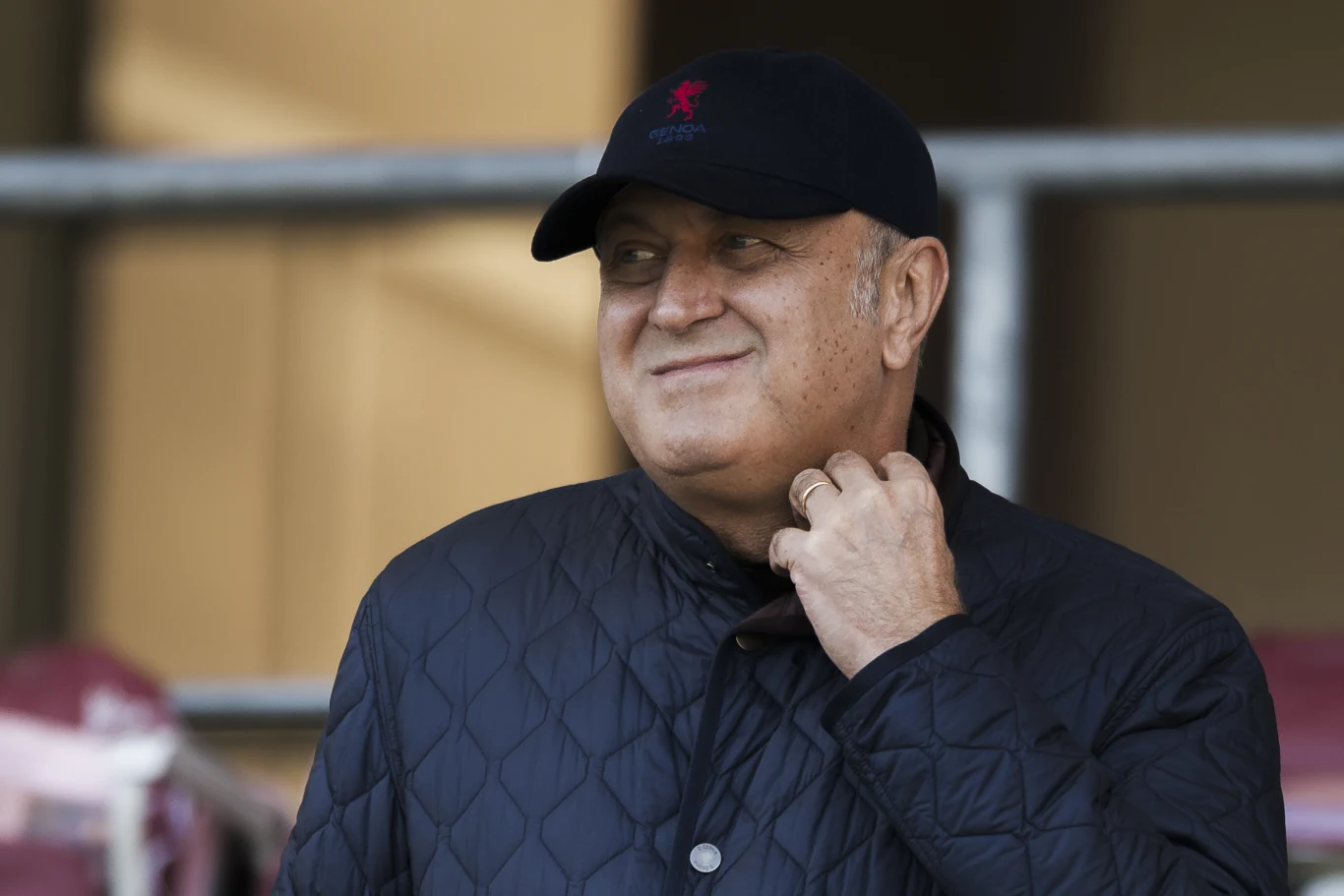 Mężczyzna w czapce z daszkiem i pikowanej kurtce uśmiecha się, trzymając rękę przy szyi, w tle rozmyte elementy infrastruktury.