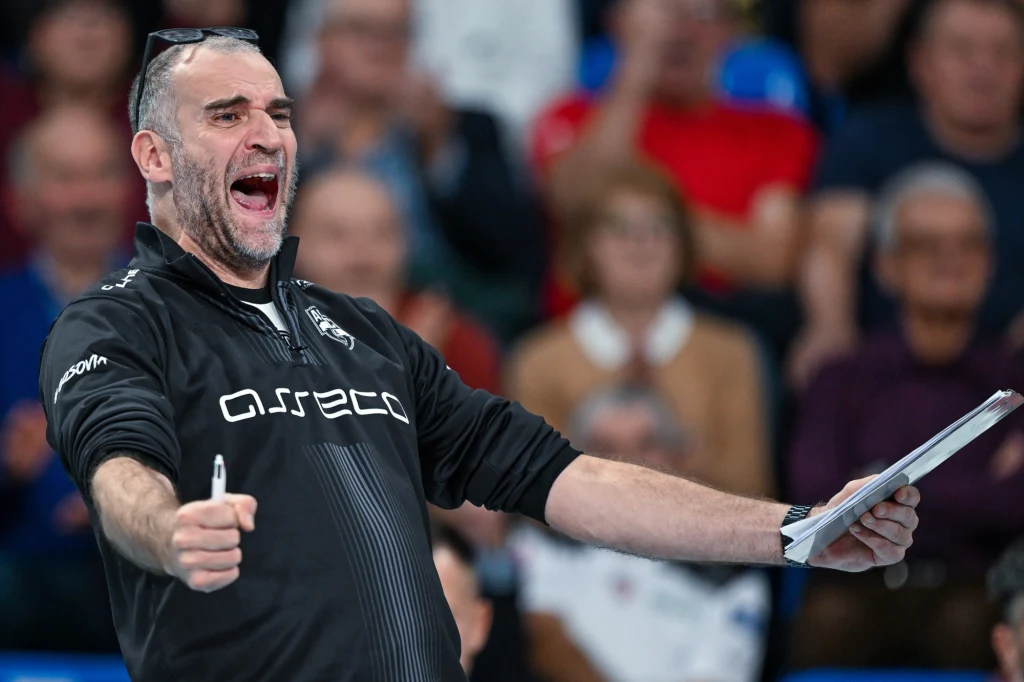 Massimo Botti, trener Asseco Resovii Trener drużyny sportowej w emocjonalnym geście, ubrany w czarną bluzę z logo Asseco, trzyma kartkę oraz długopis. W tle widownia wypełniona kibicami obserwującymi wydarzenie.