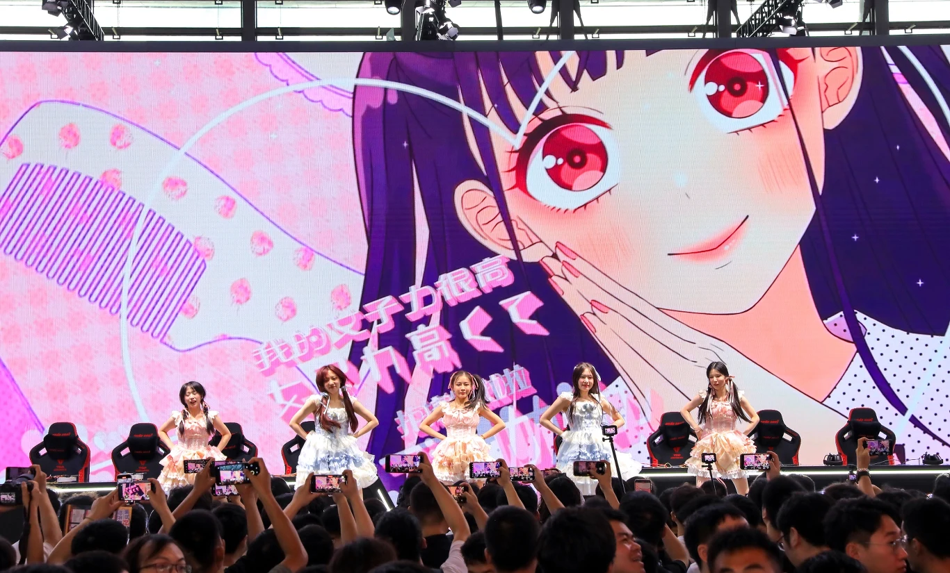 Innowacyjna terapia anime wspiera Japończyków Pięć młodych kobiet tańczy i śpiewa na scenie przed dużą publicznością, w tle wyświetlony jest ogromny rysunek postaci z anime z dużymi różowymi oczami, a widzowie trzymają telefony, nagrywając występ.