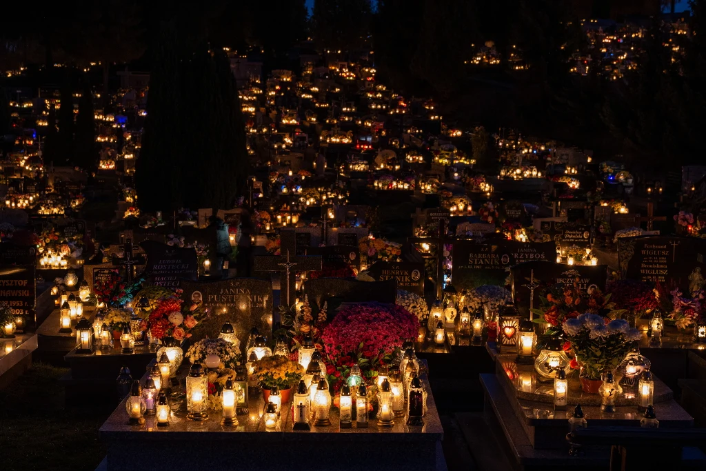 Groby oświetlone tysiącami zapalonych zniczy i otoczone kwiatami w nocnej scenerii, cisza i atmosfera zadumy na cmentarzu.