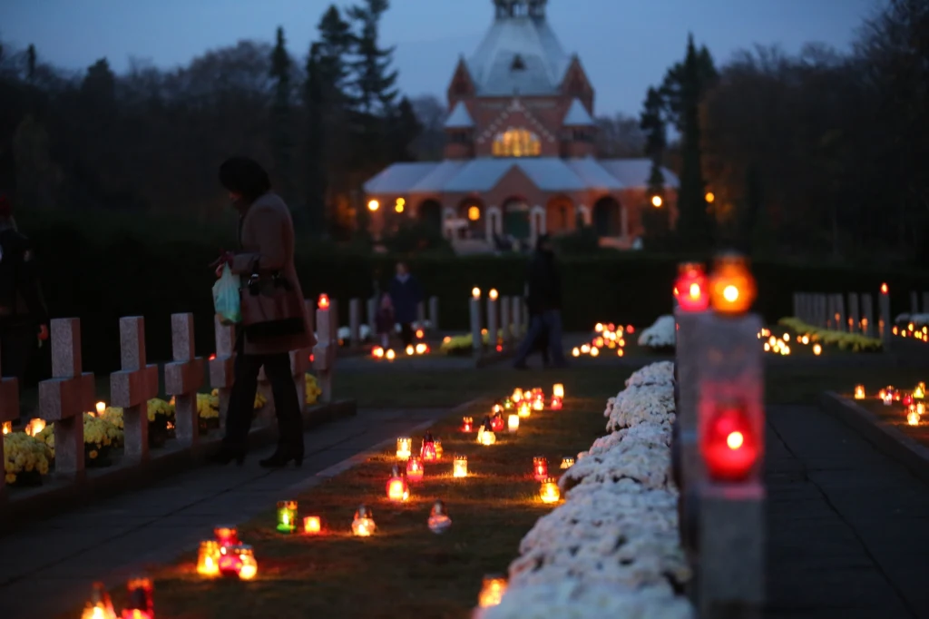 Rzędy nagrobków oświetlone licznymi płonącymi zniczami oraz kwiatami, pośród których przechadzają się ludzie, w tle widoczna kaplica na cmentarzu po zmroku.