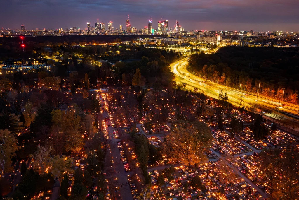 Widok na oświetlony cmentarz wśród drzew, mnóstwo zapalonych zniczy tworzy ciepłą poświatę. W tle panorama miasta ze świecącymi budynkami i szeroka autostrada rozświetlona światłami samochodów, wszystko w delikatnej, wieczornej scenerii.