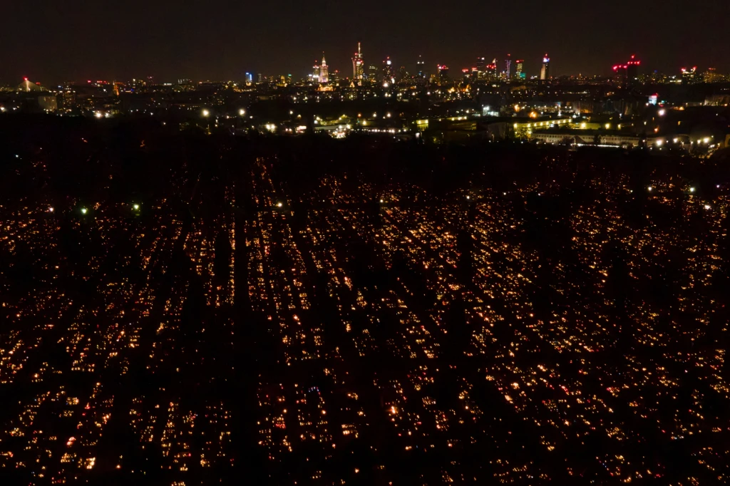 Cmentarz nocą rozświetlony tysiącami zniczy, w tle panorama rozświetlonego miasta na horyzoncie.