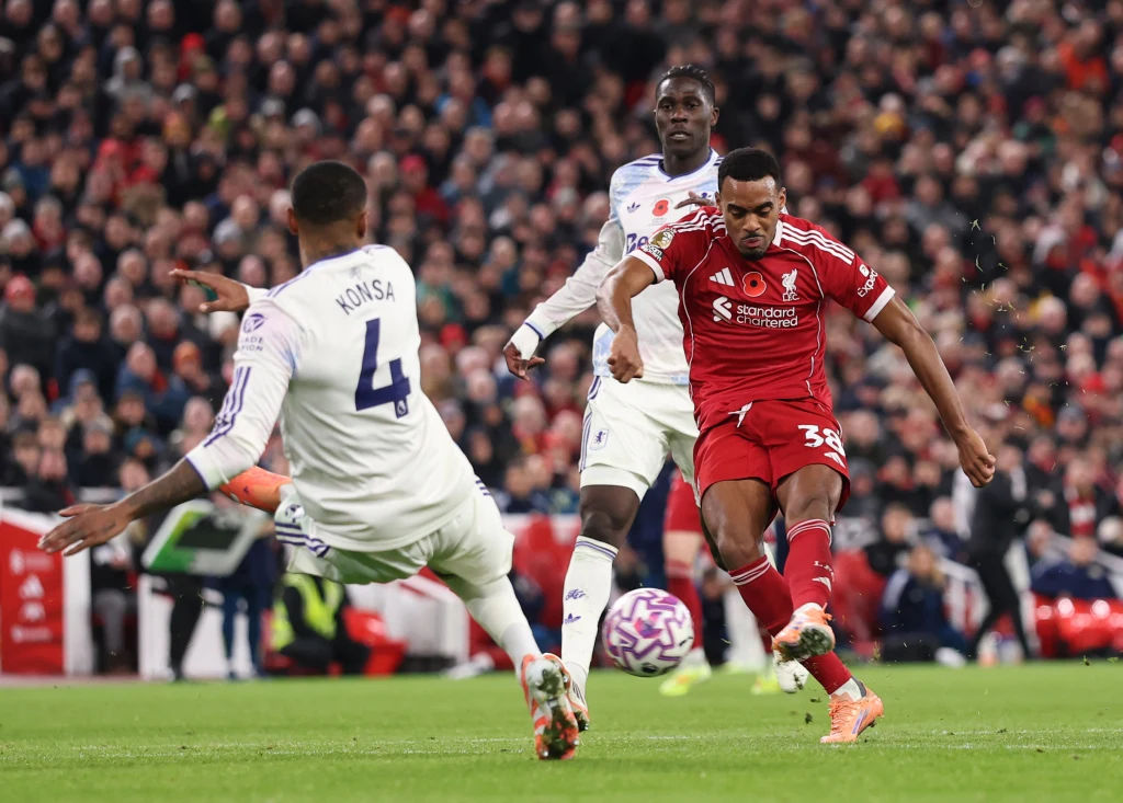 Kadr z meczu Liverpool - Aston Villa Piłkarz w czerwonym stroju wykonuje strzał na bramkę podczas meczu, otoczony przez obrońców drużyny przeciwnej na tle licznie zgromadzonej publiczności.