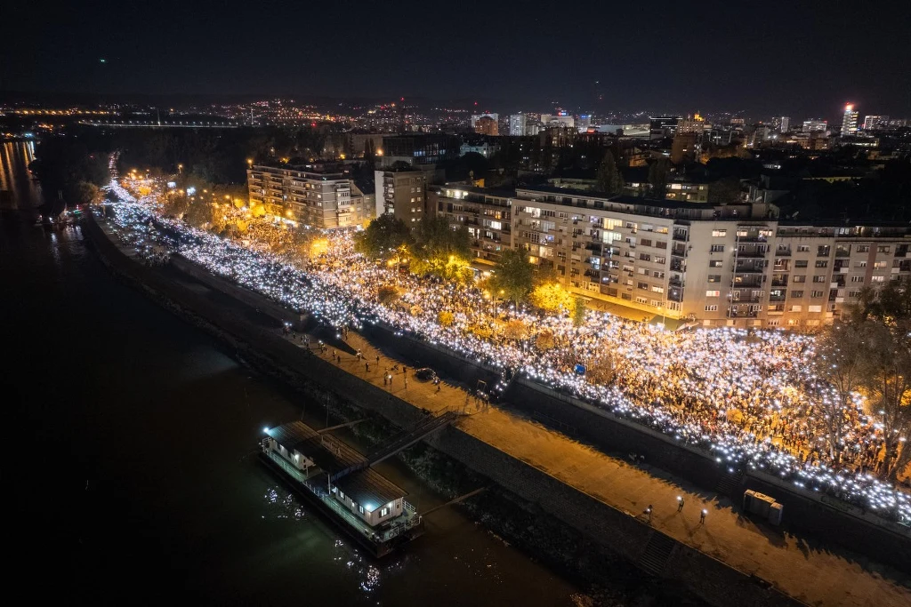 Tłum ludzi zgromadzony nocą wzdłuż nabrzeża miejskiego, setki osób oświetlają okolicę światłami swoich telefonów, wokół widoczne są wysokie budynki mieszkalne i łodzie przycumowane do brzegu.