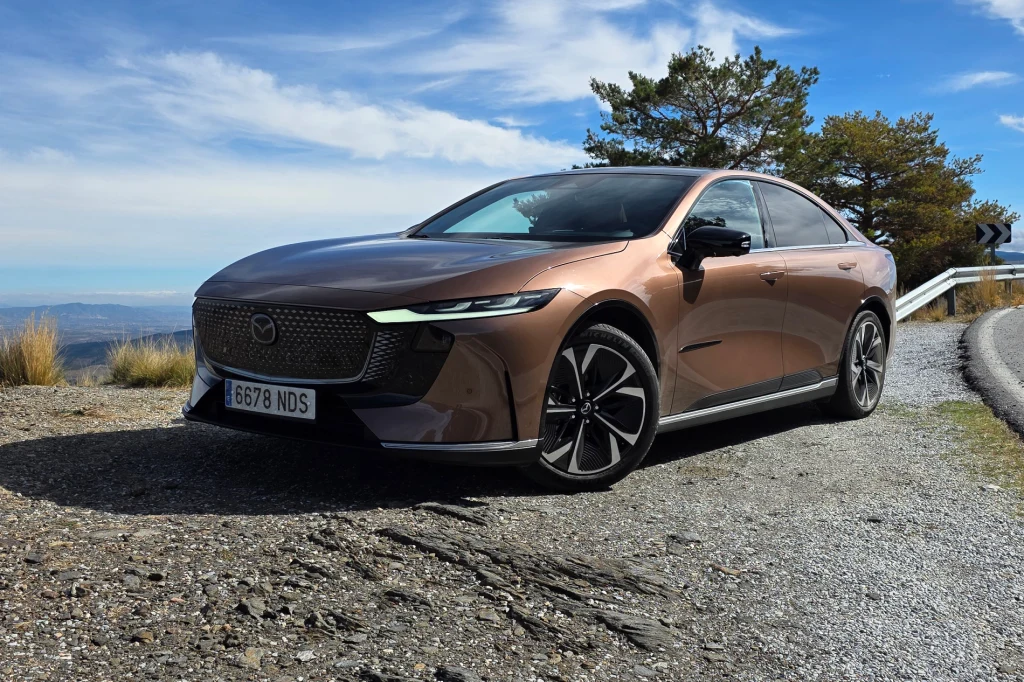 Mazda 6e może “sprzedać się” wyglądem, komfortem i niskim zużyciem energii - z praktycznego punktu widzenia trudno jednak wróżyć sukces wersji Long Range, która jest droższa, a maksymalna moc ładowania akumulatorów wynosi jedynie 90 kW. Brązowy samochód osobowy marki Mazda zaparkowany na żwirowym poboczu z widokiem na pagórkowaty krajobraz, pod jasnym, lekko zachmurzonym niebem oraz z widocznymi drzewami w tle.