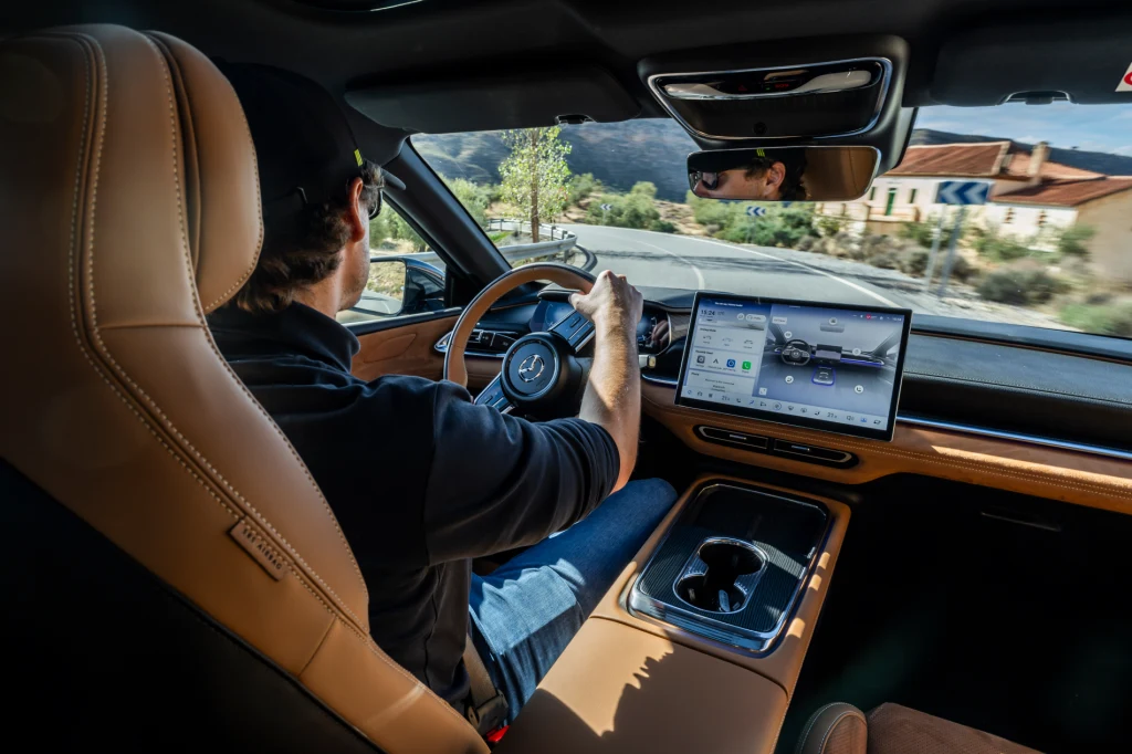 Wygodne fotele i wysoka pozycja za kierownicą plus dobrej jakości materiały - szczególne wrażenie robią zamszowe obicia. Kierowca w czapce i okularach prowadzi nowoczesny samochód z jasnym, skórzanym wnętrzem, duży ekran dotykowy na konsoli centralnej wyświetla informacje dotyczące nawigacji, za oknem widoczny zakręt drogi oraz budynki i góry w tle.