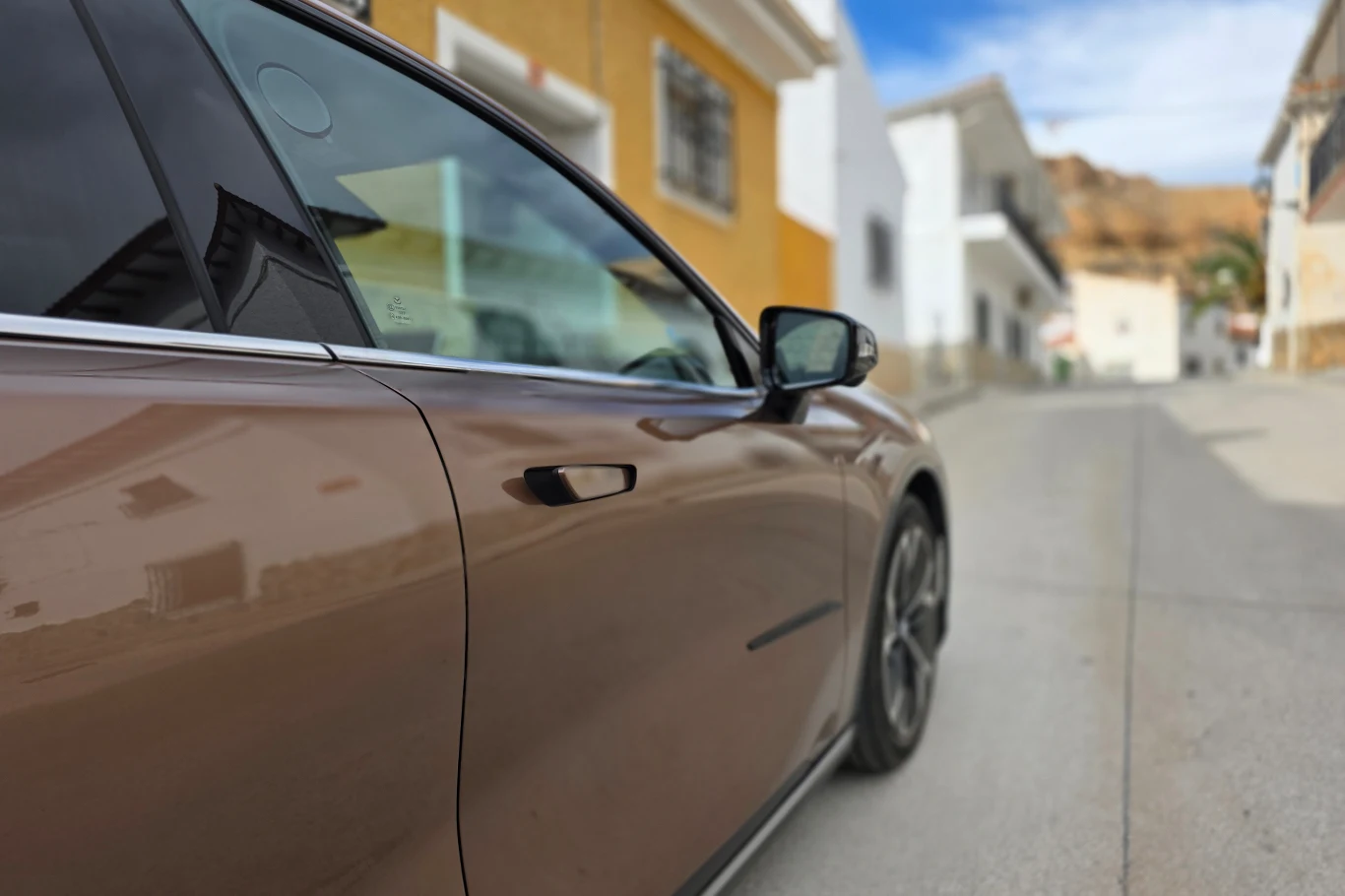 Wysuwane klamki zostaną zakazane Brązowy samochód parkujący przy ulicy, widoczny fragment bocznej karoserii oraz lusterko, w tle rozmyte budynki mieszkalne w jasnych kolorach.