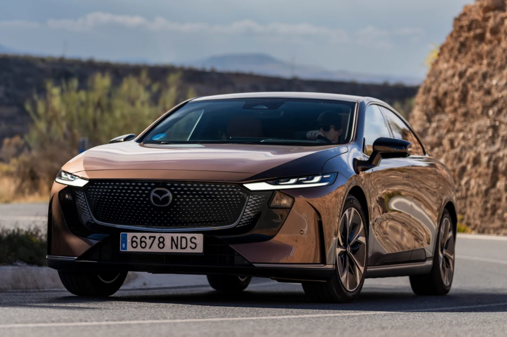 Nowa Mazda 6e to pierwszy model elektryczny Mazdy, który ma realne szanse na rynku. Jego główny rywal to Tesla Model 3 Brązowy samochód marki Mazda jedzie po asfaltowej drodze w słoneczny dzień, w tle widoczna skała oraz roślinność.