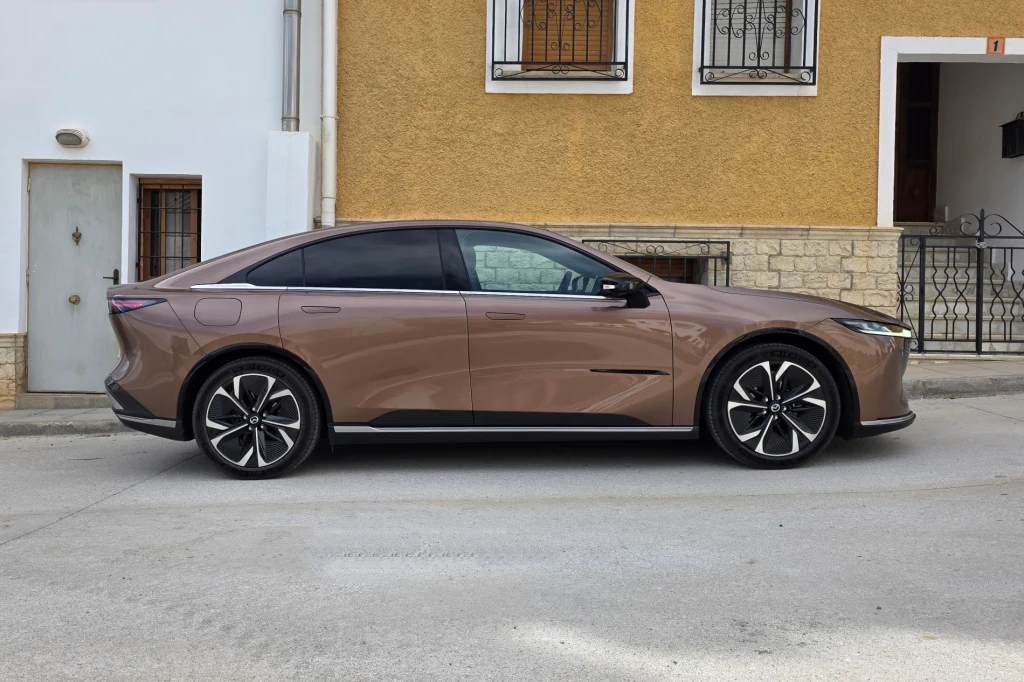 Mazda 6e to kawał auta - na długość mierzy ponad 4,9 m, czyli wyraźnie więcej niż Tesla Model 3 Brązowy, nowoczesny samochód o sportowej sylwetce zaparkowany przy jasnej elewacji budynku z żółtą i białą fasadą, ozdobioną metalowymi kratami na oknach.