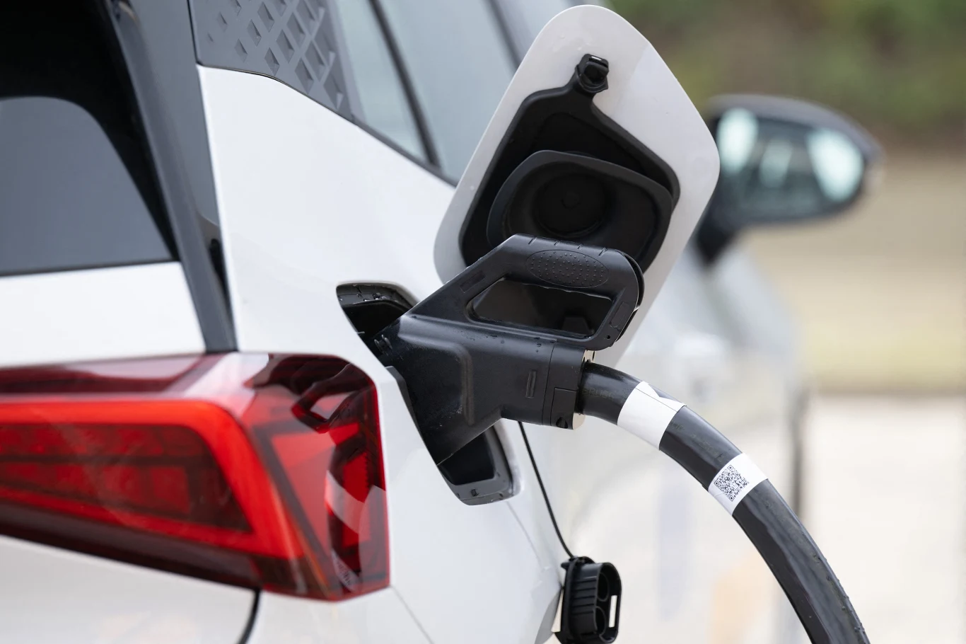 Ładowarka podłączona do portu ładowania białego samochodu elektrycznego, widoczne zbliżenie tylnej części pojazdu wraz z otwartą klapką wlewu.