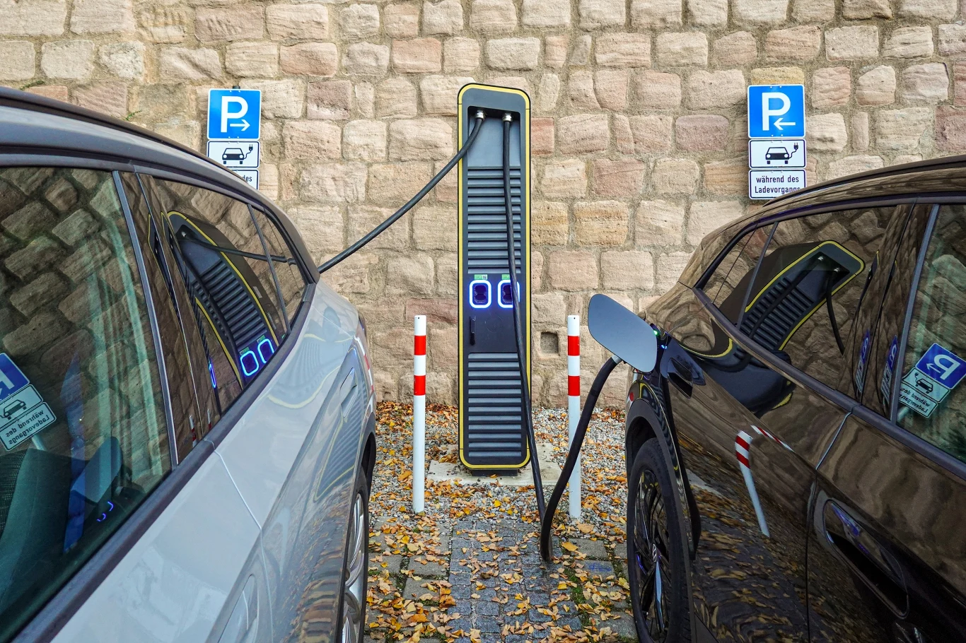 Od 2027 r. obowiązkowy będzie paszport baterii zawierający najważniejsze informacje, w tym o stanie akumulatora Stacja ładowania dla samochodów elektrycznych znajdująca się pomiędzy dwoma zaparkowanymi autami, na tle kamiennego muru. Opadłe liście na ziemi, znaki parkingowe wskazujące miejsce dla pojazdów elektrycznych.