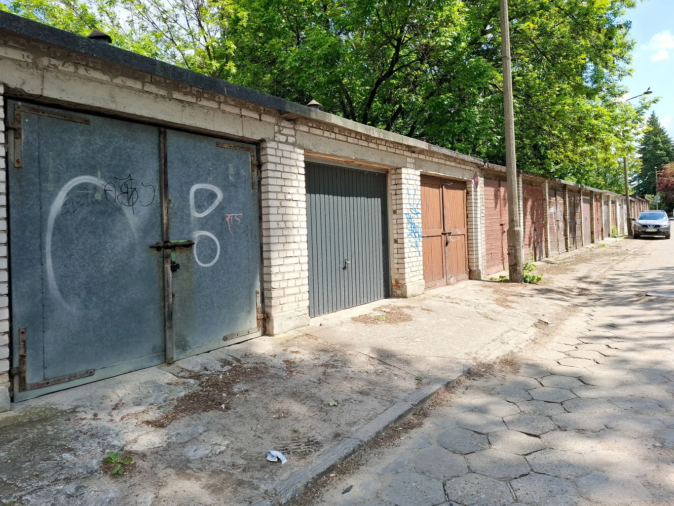 Opodatkowanie garaży wciąż budzi kontrowersje. RPO ma jasne stanowisko w tej sprawie. Zdj. ilustracyjne Rząd murowanych garaży z metalowymi drzwiami, część z nich pomalowana lub pokryta graffiti, obok widać wybrukowaną uliczkę, latarnię oraz zaparkowany na końcu biały samochód, drzewa w tle tworzą zacienioną atmosferę.